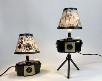 May include: Two vintage camera lamps with black and white patterned lampshades. The lamps are made from vintage cameras with a tripod stand. One lampshade has a design of a woman in a dress and the other has a design of a man in a suit.