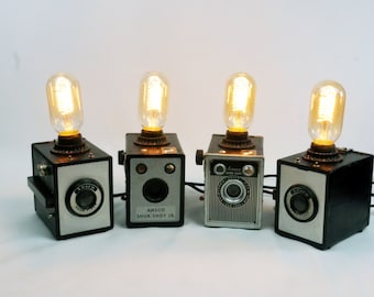 Puede incluir: Cuatro lámparas vintage con forma de cámara con bombillas encendidas. Las lámparas son negras con detalles plateados y tienen el texto "Ansco Shur-Shot Jr." en una de las lámparas.
