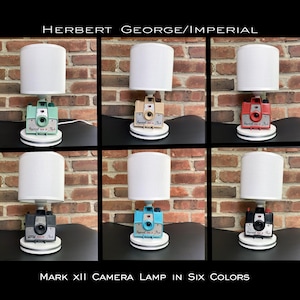 May include: Six vintage camera lamps with white lampshades. The cameras are painted in different colors: teal, beige, red, blue, black, and white. The lamps are labeled "Mark XII Camera Lamp in Six Colors".
