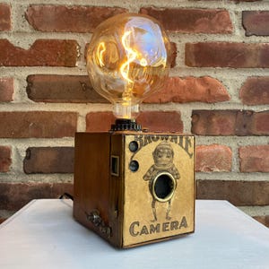 May include: A vintage-style table lamp made from a repurposed Brownie camera. The lamp features a clear glass bulb with a filament that resembles a spiral. The camera is painted brown and has a label that reads "Brownie Camera".