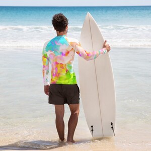 K&ouml;nnte beinhalten: Eine Person steht am Strand und h&auml;lt ein wei&szlig;es Surfbrett. Sie tr&auml;gt ein lang&auml;rmliges Shirt mit einem farbenfrohen abstrakten Muster und schwarze Shorts.