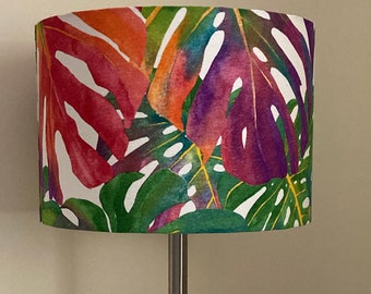 May include: A silver table lamp with a white lampshade featuring a colorful tropical leaf pattern. The lampshade has a watercolor effect with shades of pink, purple, green, and orange.