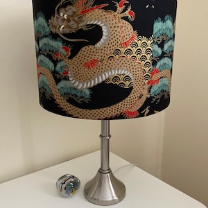 May include: A silver table lamp with a black drum lampshade featuring a gold and red dragon design. The lampshade has a pattern of pine trees and waves.