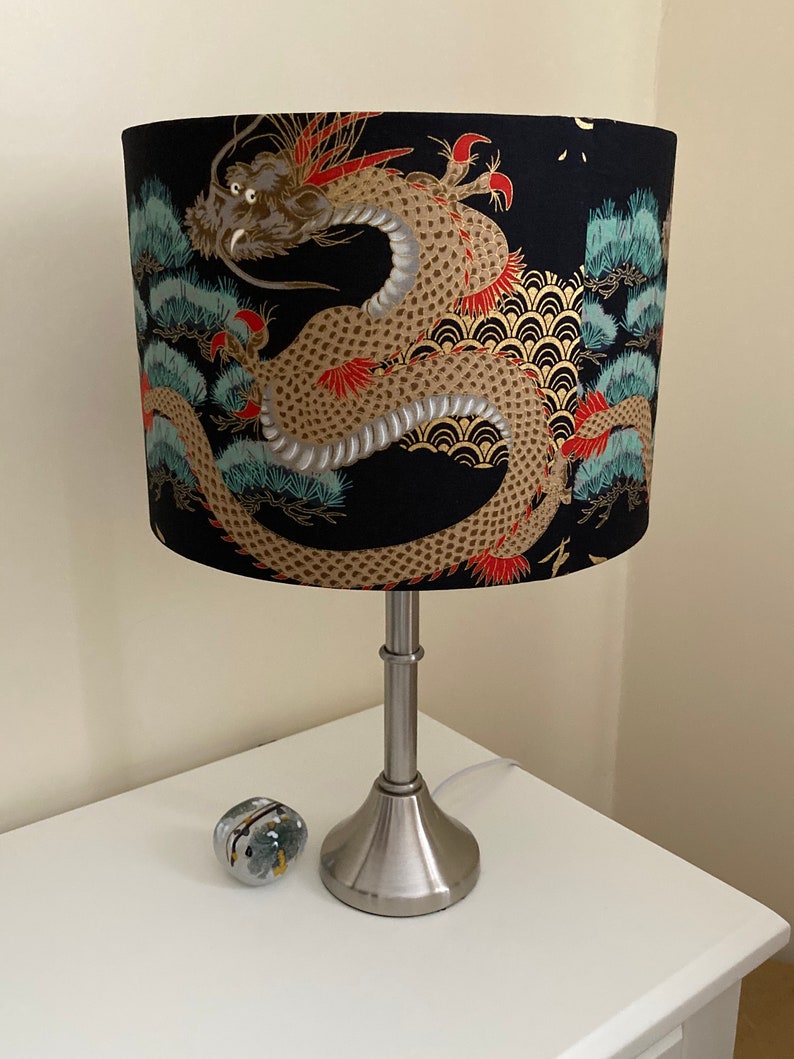 May include: A silver table lamp with a black drum lampshade featuring a gold and red dragon design. The lampshade has a pattern of pine trees and waves.