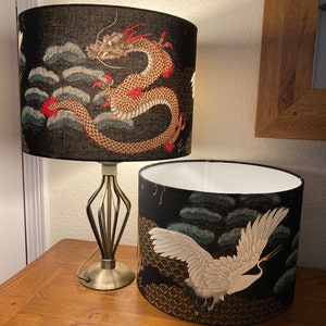 May include: Two black lampshades with a gold and white design. The top lampshade features a red and gold dragon, while the bottom lampshade features a white crane flying over a gold and black background.
