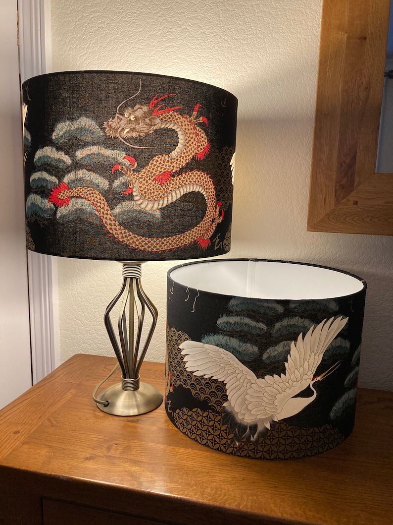 May include: Two black lampshades with a gold and white design. The top lampshade features a red and gold dragon, while the bottom lampshade features a white crane flying over a gold and black background.