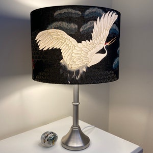 May include: A silver table lamp with a black and white crane patterned lampshade. The lampshade has a dark blue background with a white crane flying across it. The crane has a red beak and white feathers.