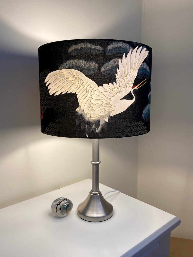 May include: A silver table lamp with a black and white crane patterned lampshade. The lampshade has a dark blue background with a white crane flying across it. The crane has a red beak and white feathers.