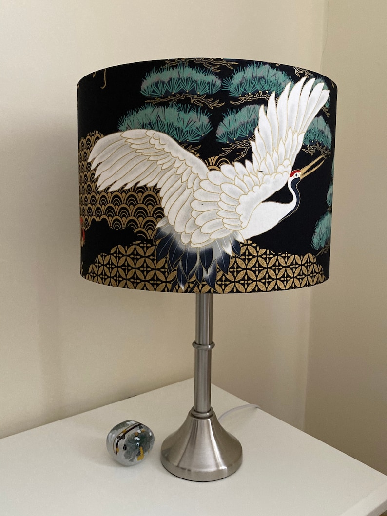 May include: A black and gold table lamp with a white crane and pine tree design on the lampshade.