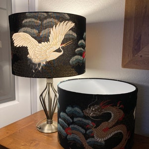 May include: Two lampshades with black backgrounds and gold accents. One lampshade features a white crane flying over a blue and green landscape. The other lampshade features a gold dragon with red accents over a blue and green landscape.