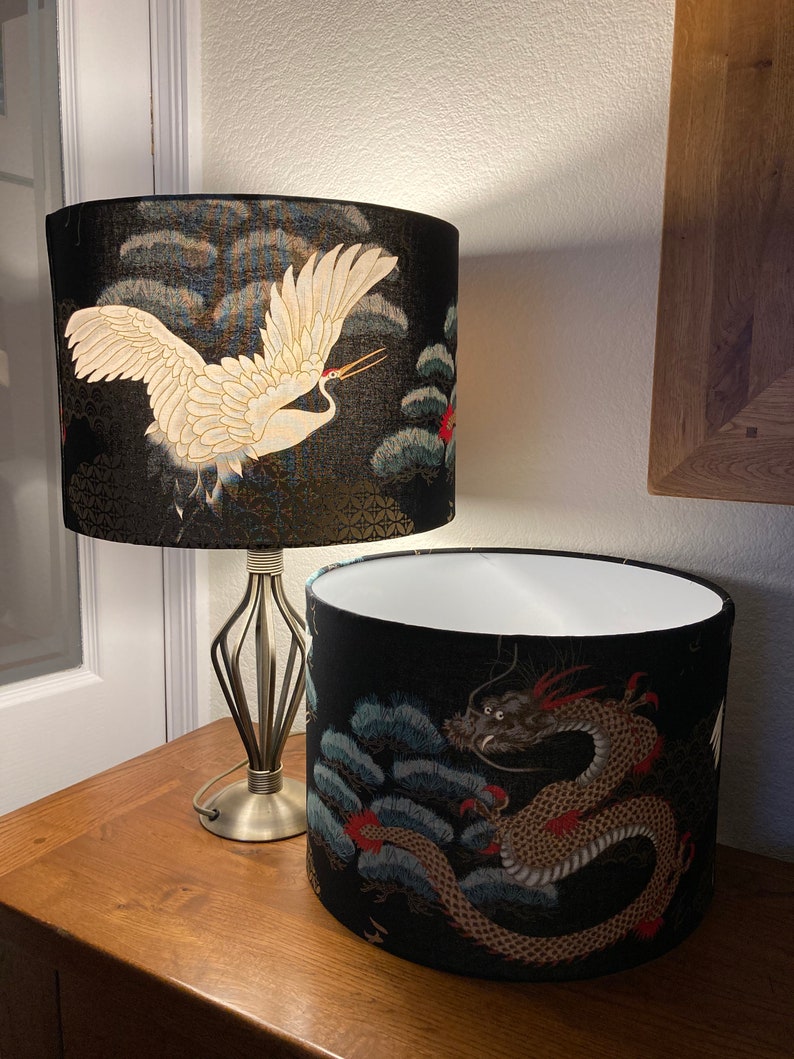 May include: Two lampshades with black backgrounds and gold accents. One lampshade features a white crane flying over a blue and green landscape. The other lampshade features a gold dragon with red accents over a blue and green landscape.