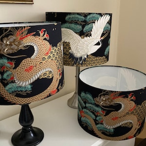 May include: Three lampshades with black backgrounds and gold accents. One lampshade features a white crane with outstretched wings, while the other two feature a gold dragon with red accents. The dragon lampshades have a blue and green pine tree design.