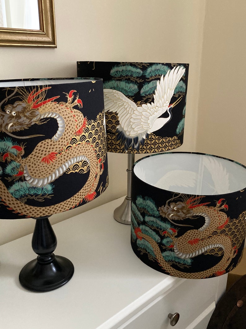 May include: Three lampshades with black backgrounds and gold accents. One lampshade features a white crane with outstretched wings, while the other two feature a gold dragon with red accents. The dragon lampshades have a blue and green pine tree design.