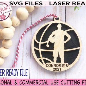 May include: A laser-cut wooden basketball ornament with a player silhouette, the name "Connor," and the number "18" and the year "2021." The ornament is round with a basketball design. The text "LASER READY FILE" is also visible.