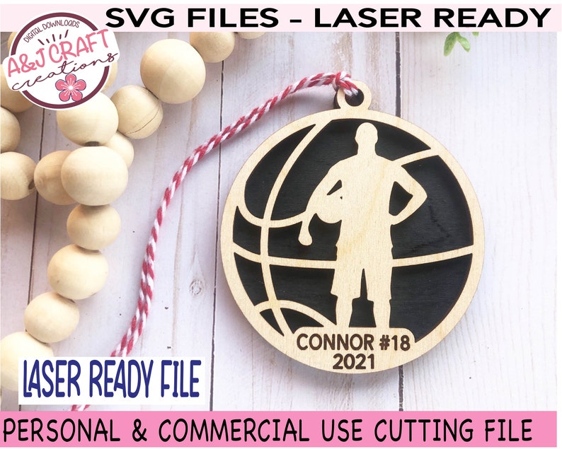 May include: A laser-cut wooden basketball ornament with a player silhouette, the name "Connor," and the number "18" and the year "2021." The ornament is round with a basketball design. The text "LASER READY FILE" is also visible.