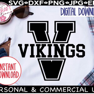 May include: A white t-shirt with a black graphic of a large letter V with the word "VIKINGS" in black text below it. The graphic is outlined in white. The t-shirt is on a wooden surface with a pair of sunglasses, a pair of jeans, and a plaid shirt.