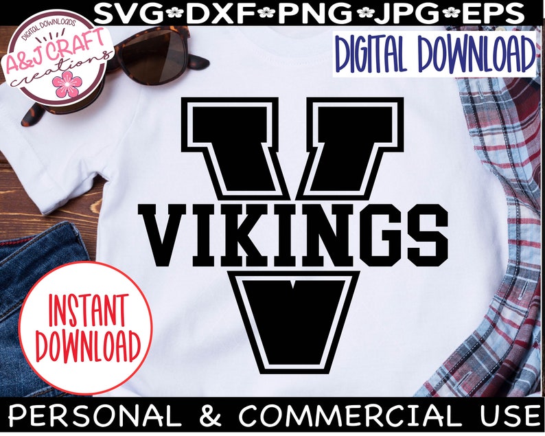 May include: A white t-shirt with a black graphic of a large letter V with the word "VIKINGS" in black text below it. The graphic is outlined in white. The t-shirt is on a wooden surface with a pair of sunglasses, a pair of jeans, and a plaid shirt.