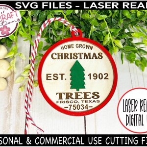 Op de afbeelding: Een rood-wit koord hangt aan een ronde houten ornament met de tekst "Home Grown Christmas Est. 1902 Trees Frisco, Texas -75034-." De ornament is laser-ready om te snijden.