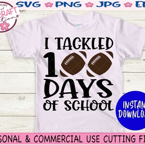May include: A light pink short-sleeve t-shirt featuring the black text I TACKLED 100 DAYS OF SCHOOL with two brown footballs replacing the zeros in 100. The design is centered on the front of the shirt, which is displayed against a light wood background.