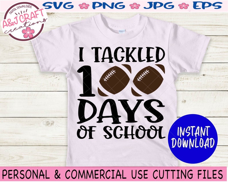 May include: A light pink short-sleeve t-shirt featuring the black text I TACKLED 100 DAYS OF SCHOOL with two brown footballs replacing the zeros in 100. The design is centered on the front of the shirt, which is displayed against a light wood background.