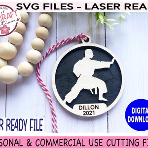 May include: A laser-cut wooden ornament featuring a silhouette of a person in a karate pose. The ornament is black with a white silhouette and the text "Dillon 2021".