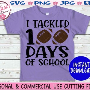 May include: A light purple t-shirt featuring the black text I TACKLED 100 DAYS OF SCHOOL with two brown football graphics replacing the zeros in 100. The design includes a circular logo in the top left corner and a blue circle in the bottom right.