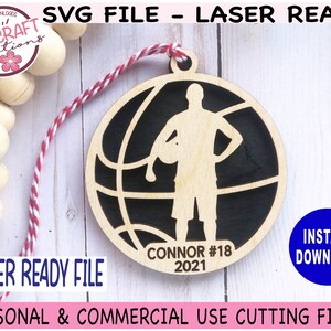 May include: A laser-ready SVG file for a basketball-themed ornament. The design features a basketball player silhouette inside a basketball, with the name "CONNOR #18 2021" at the bottom. The ornament is made of light-colored wood with a black background.