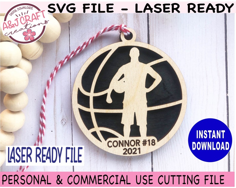 May include: A laser-ready SVG file for a basketball-themed ornament. The design features a basketball player silhouette inside a basketball, with the name "CONNOR #18 2021" at the bottom. The ornament is made of light-colored wood with a black background.