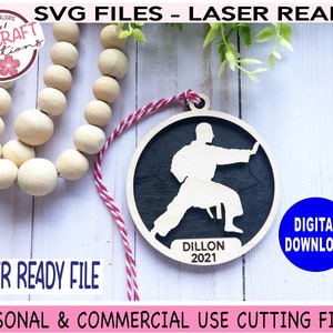 May include: A laser-cut wooden ornament featuring a silhouette of a person in a karate pose. The ornament is black and white and has the name "Dillon" and the year "2021" inscribed on it.