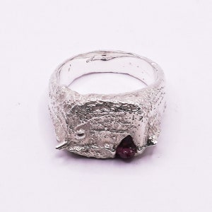 Puede incluir: Un anillo de plata con un diseño texturizado y orgánico. Una pequeña piedra preciosa de color rojo oscuro está engastada en el anillo.