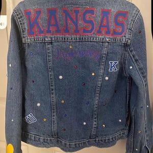 May include: A denim jacket with the word "KANSAS" in red and blue lettering across the back. The jacket is embellished with pearls, colorful gems, and patches. The name "Jayhawks" is written in cursive below the word "KANSAS".