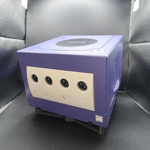 May include: A purple Nintendo GameCube console with a silver faceplate. The console has a disc tray on top and several ports on the front. The console is sitting on a black stand.