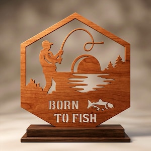 Peut inclure: Un panneau hexagonal en bois présentant une silhouette découpée au laser d'une personne pêchant avec une canne, un soleil se couchant sur l'eau et des pins. Le texte BORN TO FISH et un motif de poisson sont découpés dans la partie inférieure.