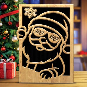 Peut inclure: Décoration de Noël en bois représentant une découpe du Père Noël portant des lunettes de soleil avec le mot "HO" sur chaque verre. Le design du Père Noël est placé sur un fond noir et encadré de bois clair. Un flocon de neige est au-dessus de la tête du Père Noël.