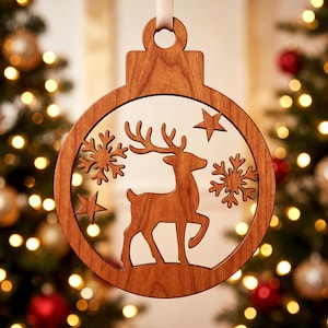 Könnte beinhalten: Ein runder Weihnachtsanhänger aus Holz mit einem lasergeschnittenen Motiv eines stehenden Rentiers in der Mitte, umgeben von zwei Schneeflocken und zwei Sternen. Der Anhänger hat eine glatte, braune Holzmaserung und eine Öse zum Aufhängen.