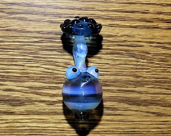 May include: A blue and black glass mushroom shaped pipe with a round body and a small, flat top. The pipe has a white stripe around the middle and two small blue eyes.