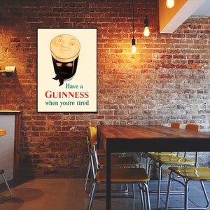 May include: A framed vintage Guinness advertisement. The poster features a smiling pint of Guinness with the text "Have a Guinness when you're tired." The poster is displayed on a brick wall in a pub setting, with a table and chairs in the foreground.