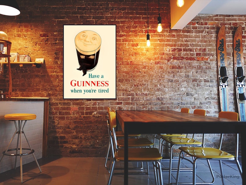 May include: A framed vintage Guinness advertisement. The poster features a smiling pint of Guinness with the text "Have a Guinness when you're tired." The poster is displayed on a brick wall in a pub setting, with a table and chairs in the foreground.