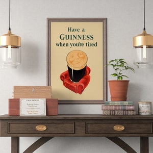 K&ouml;nnte beinhalten: Ein gerahmtes Guinness-Werbeplakat im Vintage-Stil. Das Kunstwerk zeigt ein Pint Guinness mit einem l&auml;chelnden Gesicht, das auf einem roten Sessel sitzt, mit dem Text "Have a Guinness when you're tired". Die Szene befindet sich auf einem Holztisch.
