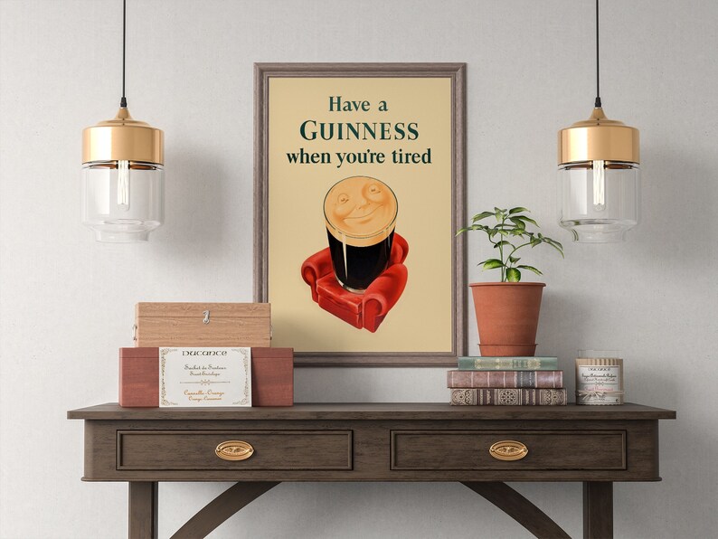 K&ouml;nnte beinhalten: Ein gerahmtes Guinness-Werbeplakat im Vintage-Stil. Das Kunstwerk zeigt ein Pint Guinness mit einem l&auml;chelnden Gesicht, das auf einem roten Sessel sitzt, mit dem Text "Have a Guinness when you're tired". Die Szene befindet sich auf einem Holztisch.