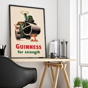 May include: A framed vintage Guinness advertisement poster. The poster features a green steamroller with the text "Guinness for strength" in red and green. The poster is displayed on a wooden desk with books and a black chair.