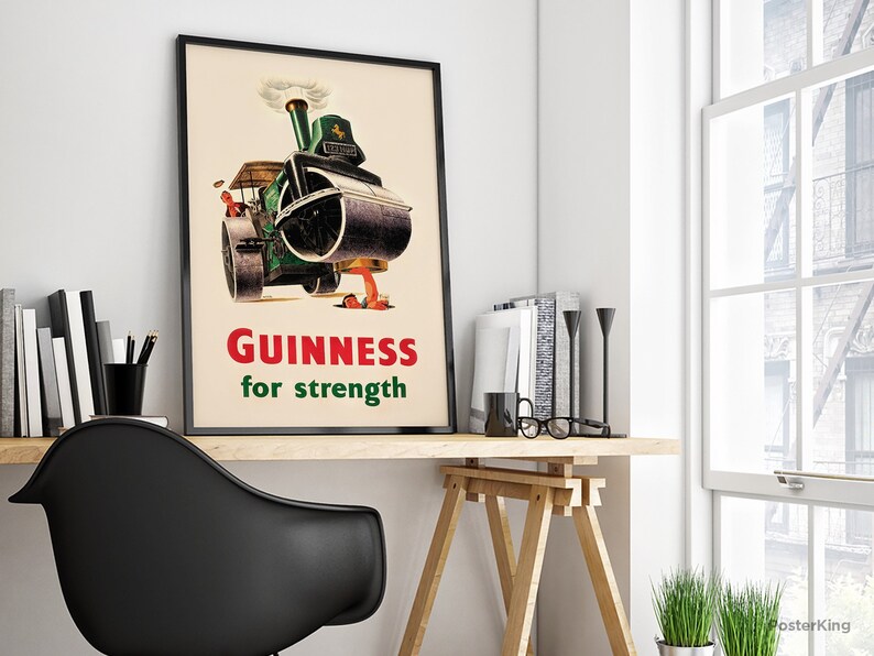 May include: A framed vintage Guinness advertisement poster. The poster features a green steamroller with the text "Guinness for strength" in red and green. The poster is displayed on a wooden desk with books and a black chair.