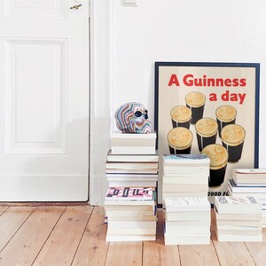 May include: A decorative skull with a rainbow pattern rests on a pile of books. Beside it is a framed poster with the text "A Guinness a day" and illustrations of smiling pints. The setting is a room with a white door and wooden floor.