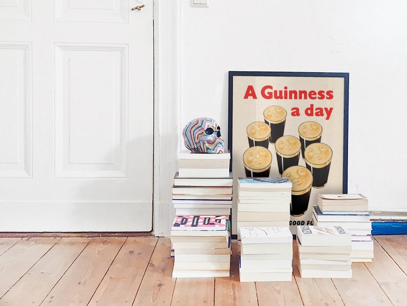 May include: A decorative skull with a rainbow pattern rests on a pile of books. Beside it is a framed poster with the text "A Guinness a day" and illustrations of smiling pints. The setting is a room with a white door and wooden floor.