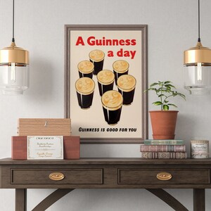 May include: A framed vintage Guinness poster with the text "A Guinness a day" in red. The poster shows smiling pints of stout. The phrase "Guinness is good for you" is at the bottom. The poster is displayed on a wooden table.