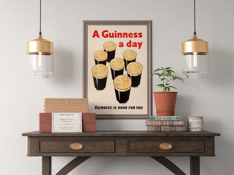 May include: A framed vintage Guinness poster with the text "A Guinness a day" in red. The poster shows smiling pints of stout. The phrase "Guinness is good for you" is at the bottom. The poster is displayed on a wooden table.