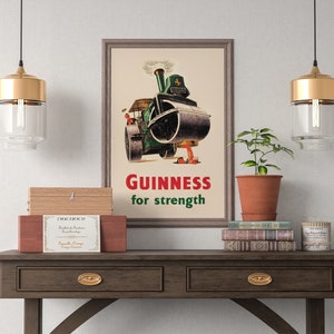 May include: Framed vintage Guinness advertisement featuring a steamroller pouring out a dark liquid. The text "Guinness for strength" is in red and green. The image is displayed on a wooden table with decorative items.