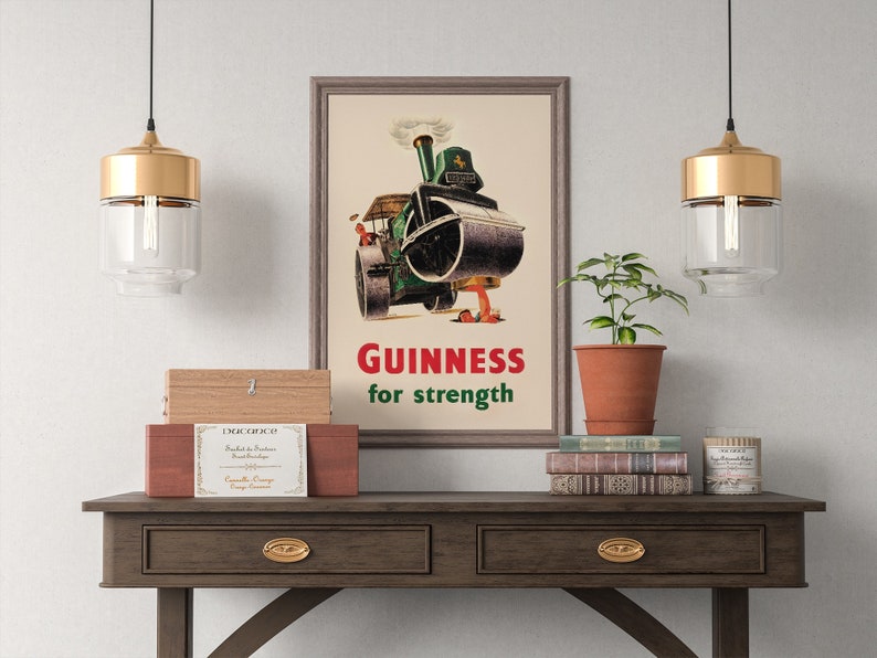 May include: Framed vintage Guinness advertisement featuring a steamroller pouring out a dark liquid. The text "Guinness for strength" is in red and green. The image is displayed on a wooden table with decorative items.