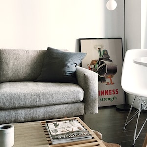 May include: A contemporary living room with a grey sofa and a dark grey cushion. A framed vintage Guinness advert is displayed. A white table and chair are in the background, along with a wooden coffee table with a magazine and a concrete candle holder.