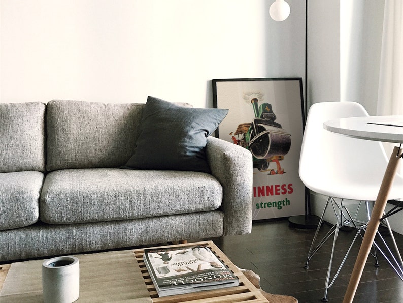 May include: A contemporary living room with a grey sofa and a dark grey cushion. A framed vintage Guinness advert is displayed. A white table and chair are in the background, along with a wooden coffee table with a magazine and a concrete candle holder.